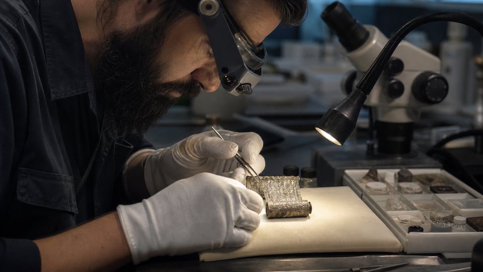 Un restaurateur déroule délicatement un minuscule rouleau d'argent sous une loupe binoculaire