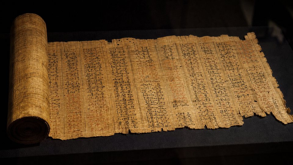 Long papyrus égyptien couvert d'écriture hiératique, partiellement déroulé dans une vitrine sombre