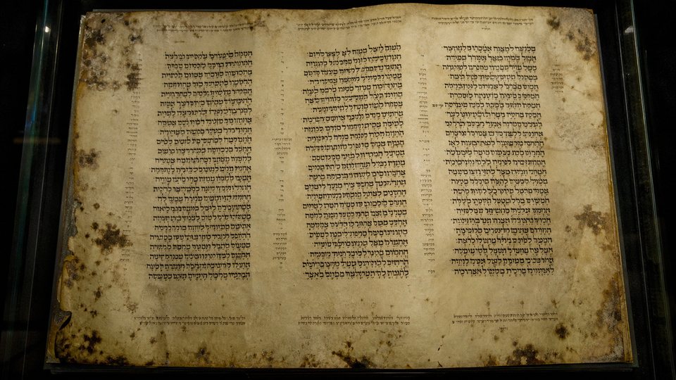 Un feuillet de parchemin taché, couvert de trois colonnes d'écriture hébraïque, exposé dans une vitrine sombre