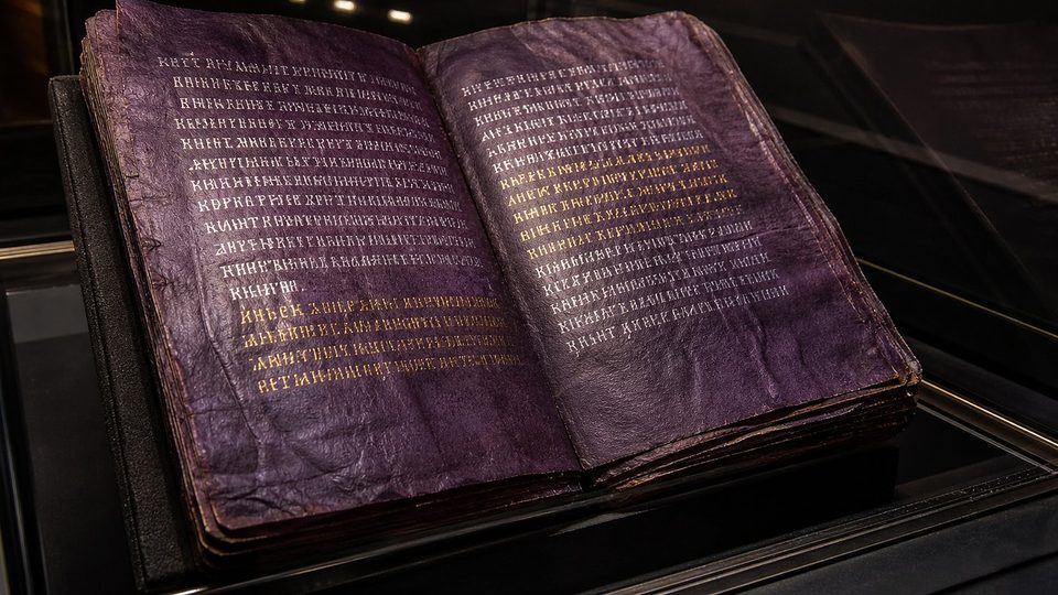 Un codex ouvert aux pages pourpres, couvertes de lettres d'argent et de quelques lignes d'or, dans une vitrine sombre