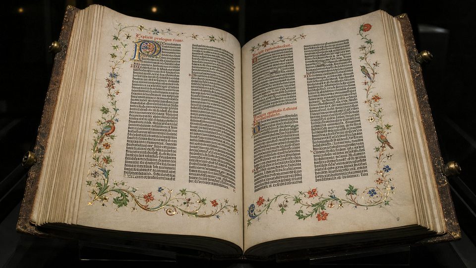 Une Bible imprimée du XVe siècle ouverte dans une vitrine : deux colonnes de texte gothique par page, titres rouges, grande initiale peinte et marges ornées de fleurs et d'oiseaux