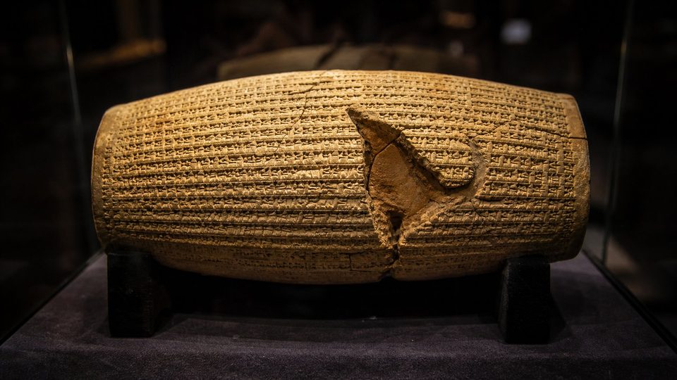 Cylindre d'argile en forme de baril couvert de fines lignes cunéiformes, posé dans une vitrine sombre